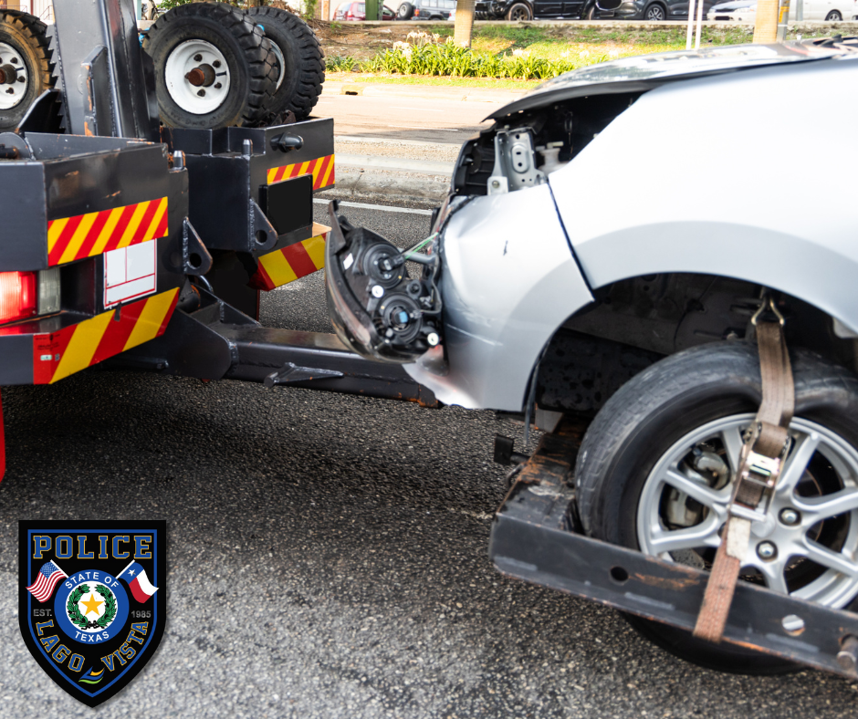 The image shows a scene involving a tow truck vehicle and a damaged car. The car being towed is silver and appears to have sustained significant damage on the driver's side door. The towing vehicle has large wheels. ther's a police badge visible in the corner, indicaating that this incident may involve law enforcement, specifically a formal relationship between law enforcement and tow services in Lago Vista, TX. 